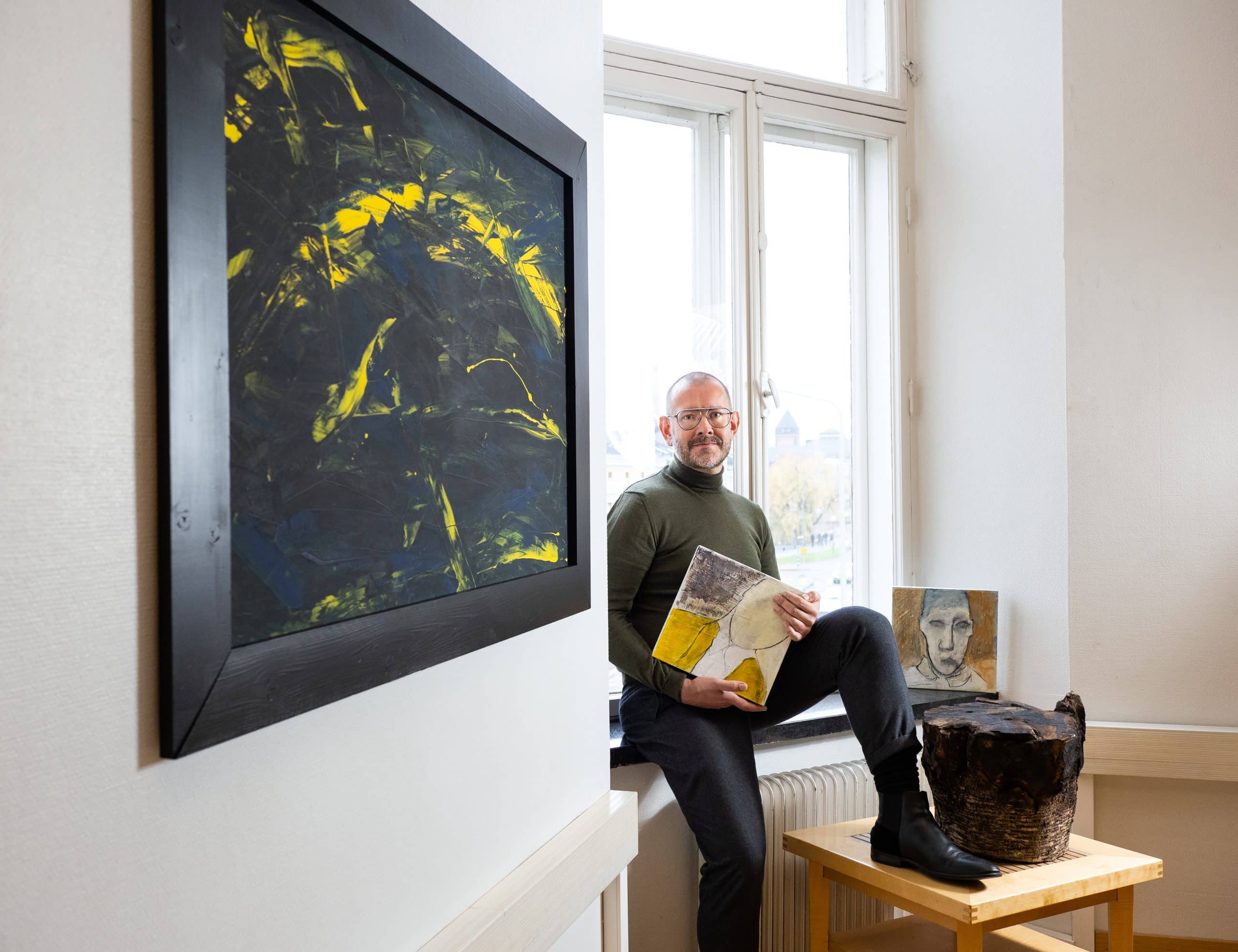 Tommi Ojala istuu ikkunan edessä maalauksia ympärillään.